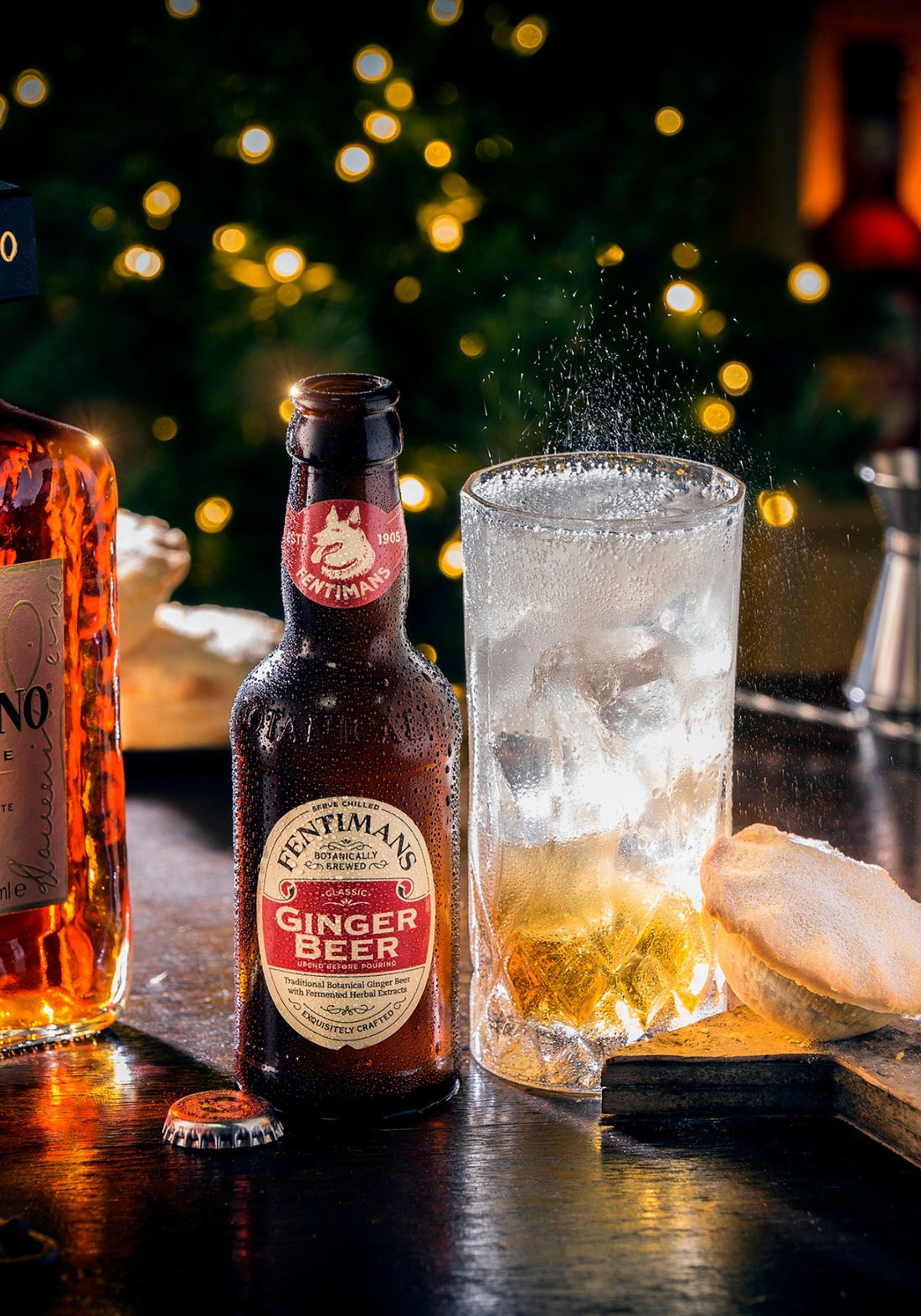A bottle of Fentimans Ginger Beer beside a highball drink in a festive setting with a mince pie on the side